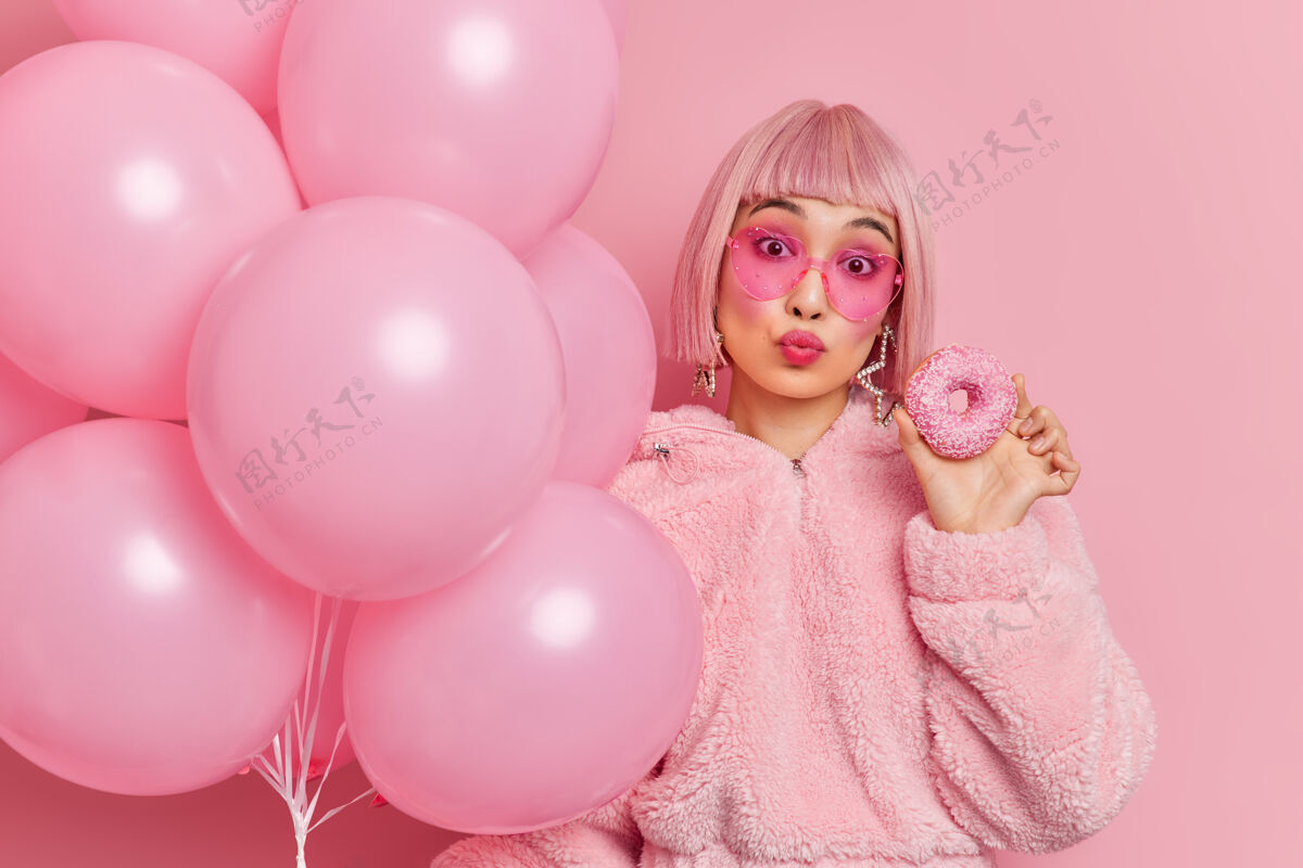 穿浪漫可爱的亚洲女人 双唇合拢 粉色波波头 穿着冬季皮衣 手拿美味的甜甜圈和充气的氦气球 在派对上和朋友庆祝生日束女性浪漫
