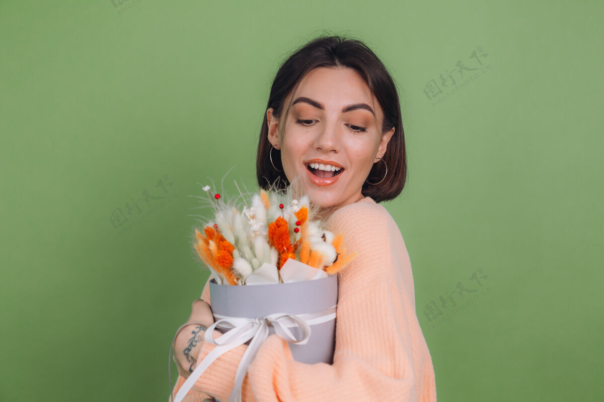 魅力年轻女子穿着休闲桃色毛衣隔离在绿橄榄墙上手持橙白色花盒组成的棉花 吉普赛拉小麦和拉古鲁斯作为礼物开心惊喜花黑发冷静