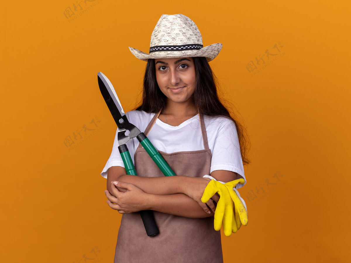 年轻穿着围裙 戴着夏帽 手持树篱剪 戴着工作手套 脸上带着微笑的年轻园丁女孩站在橙色的墙上