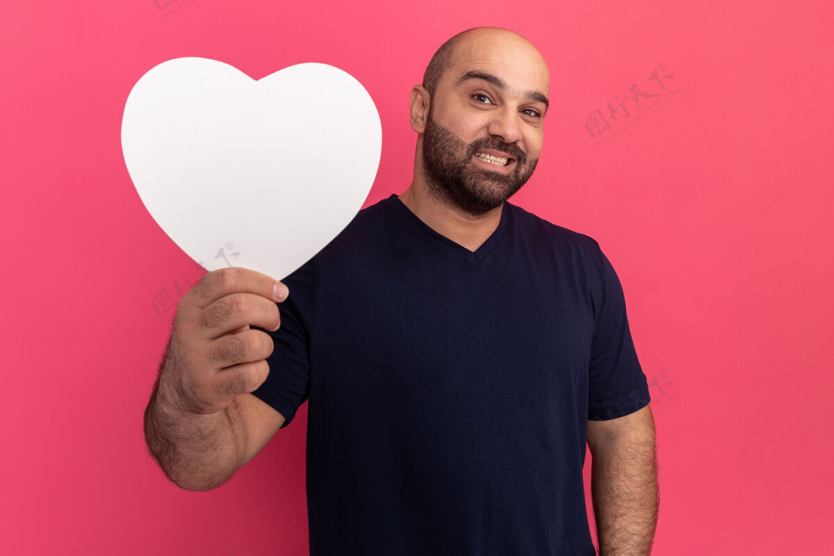 加油一个留着胡子的男人 穿着t恤 手里拿着一个硬纸板心 站在粉色的墙上开心地笑着胡子伙计站起来