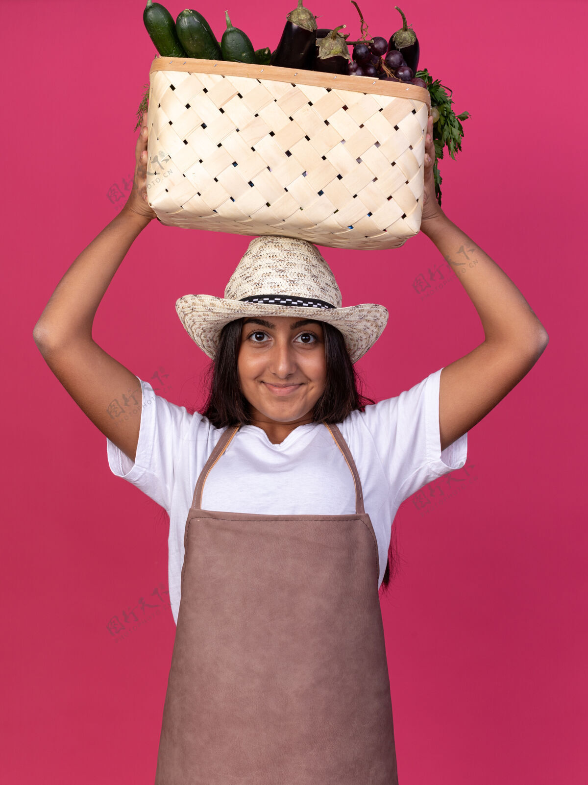 立场快乐的年轻园丁女孩 围裙戴着夏帽 头上顶着装满蔬菜的箱子 脸上带着微笑站在粉红色的墙上围裙花园帽子