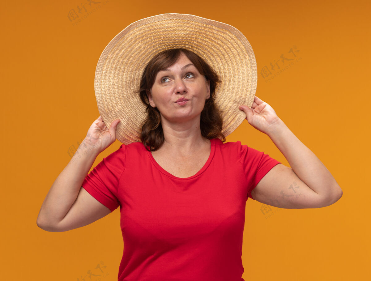 女人身穿红色t恤 头戴夏帽的中年妇女站在橘色的墙上 高兴而积极地抬头看中年立场积极