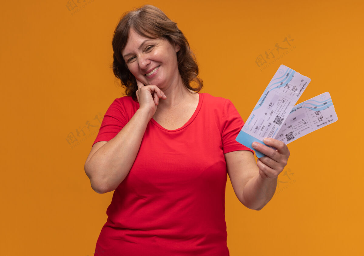 机票身穿红色t恤的中年妇女站在橙色的墙上 手里拿着一张笑脸的机票票位中年女人