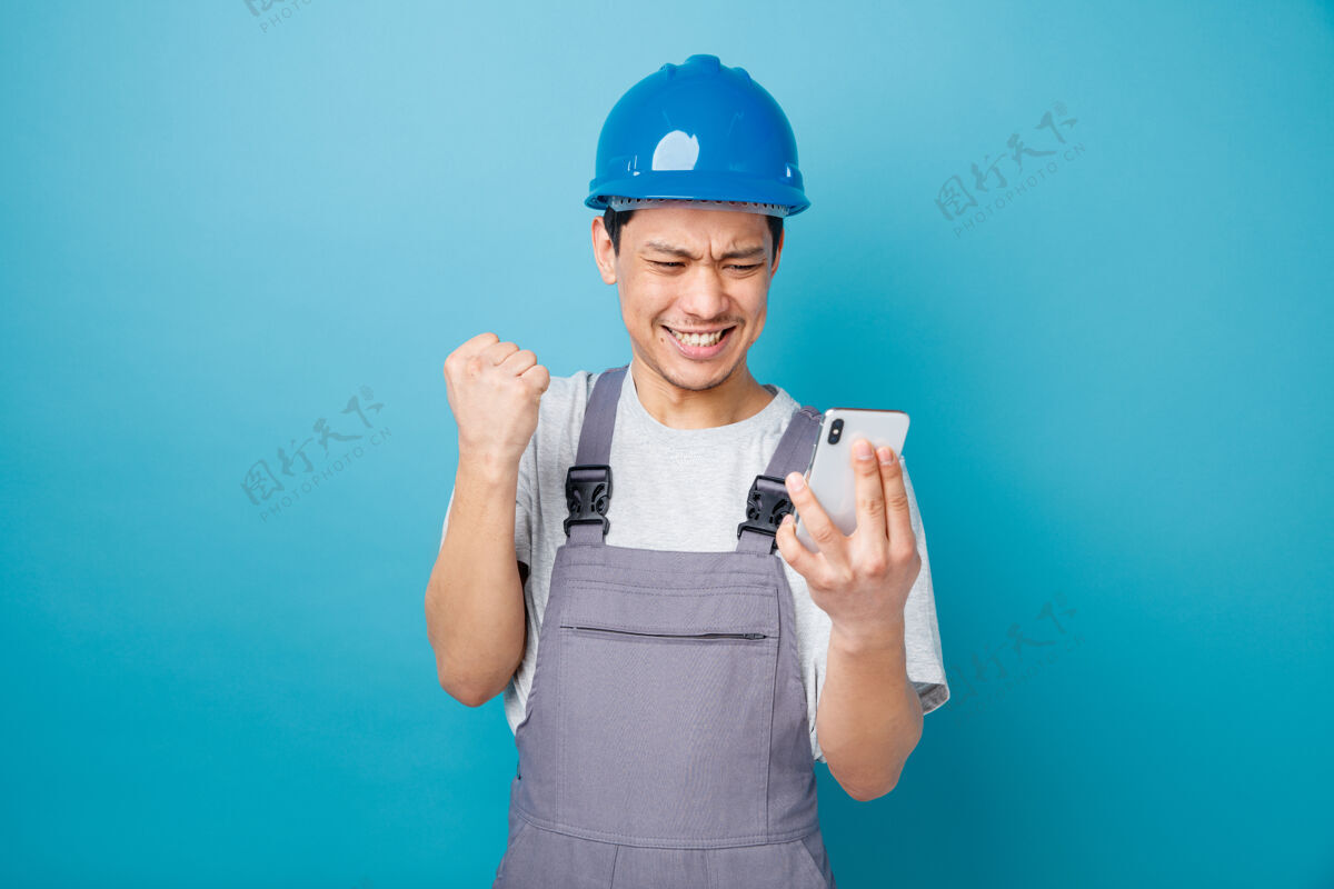 是积极进取的年轻建筑工人戴着安全帽 穿着制服 手持手机 看着手机做着“是”的手势安全电话佩戴