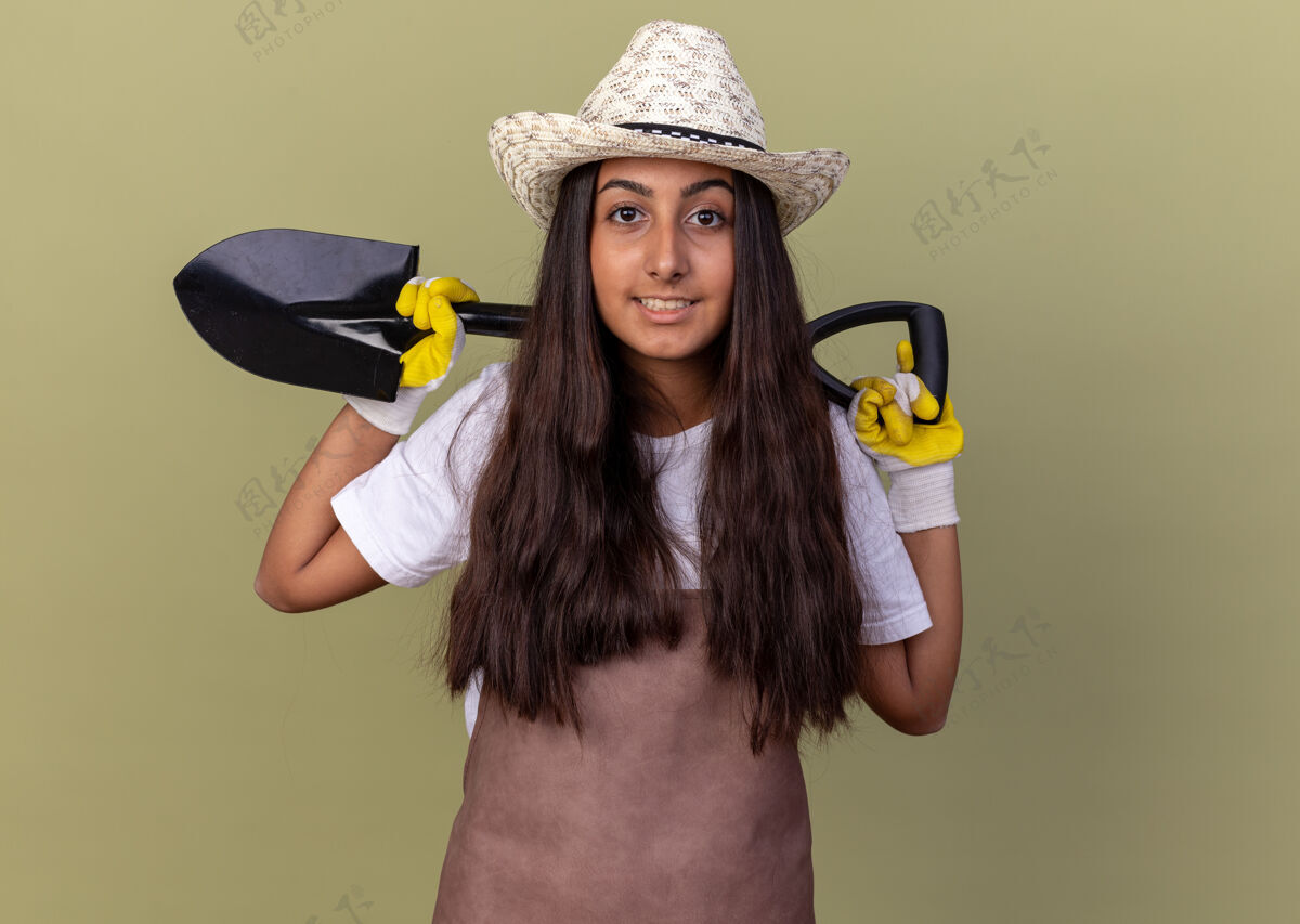 花园穿着围裙 戴着夏帽 戴着工作手套 拿着铲子 自信地站在绿色墙壁上的年轻园丁女孩帽子穿年轻的