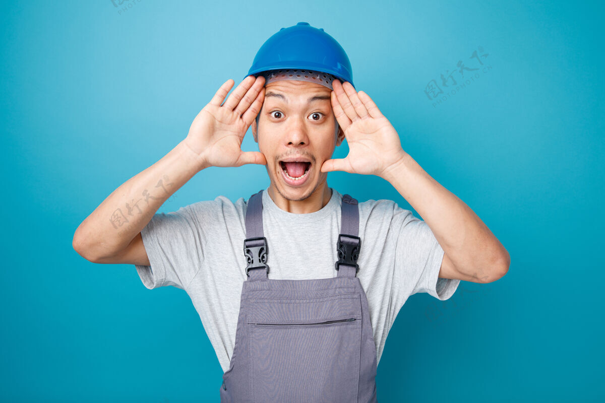 穿兴奋的年轻建筑工人戴着安全帽和制服 手举在头上保持工人头盔