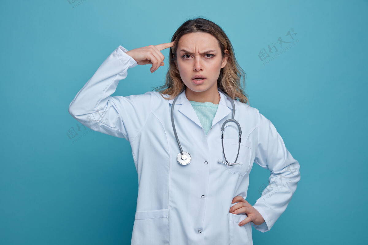 脖子困惑的年轻女医生穿着医用长袍 脖子上戴着听诊器 手放在腰上做着思考的手势腰医学穿