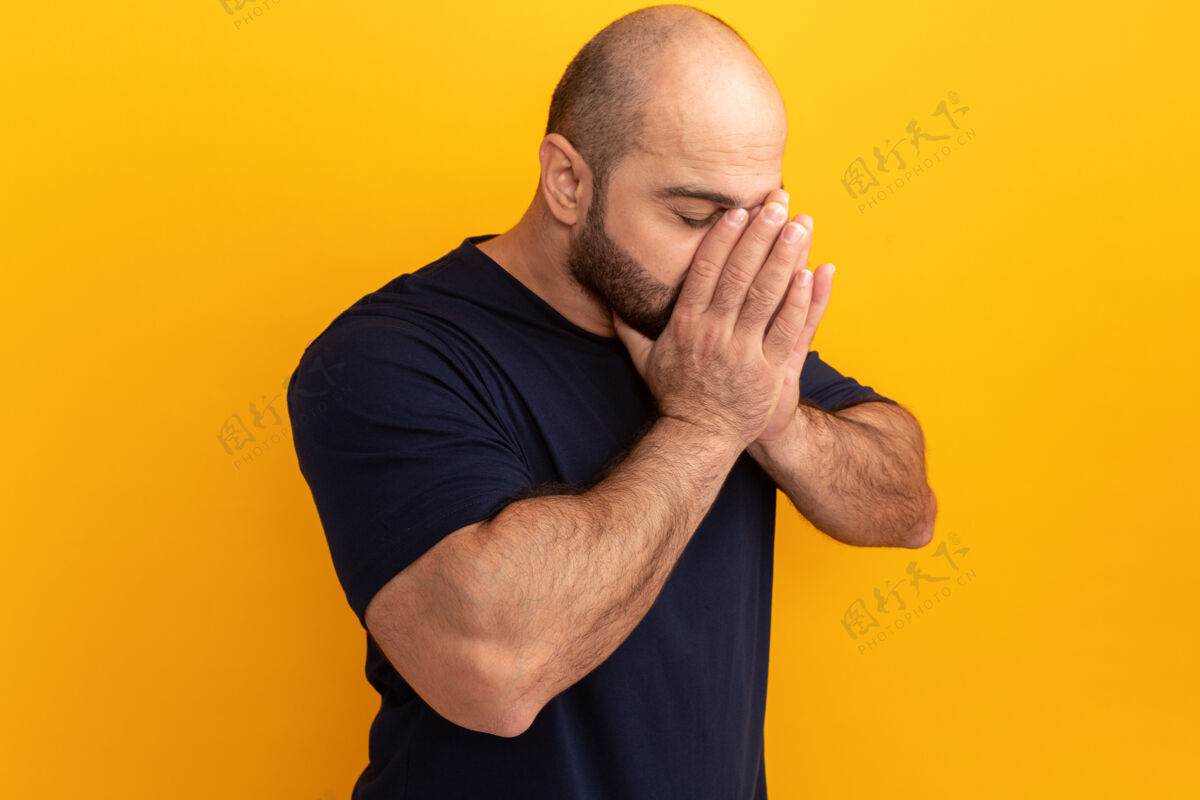 胡子一个留着大胡子的男人 穿着海军蓝t恤 手拉着手站在橘色的墙上 脸上愁眉苦脸的伙计站着担心