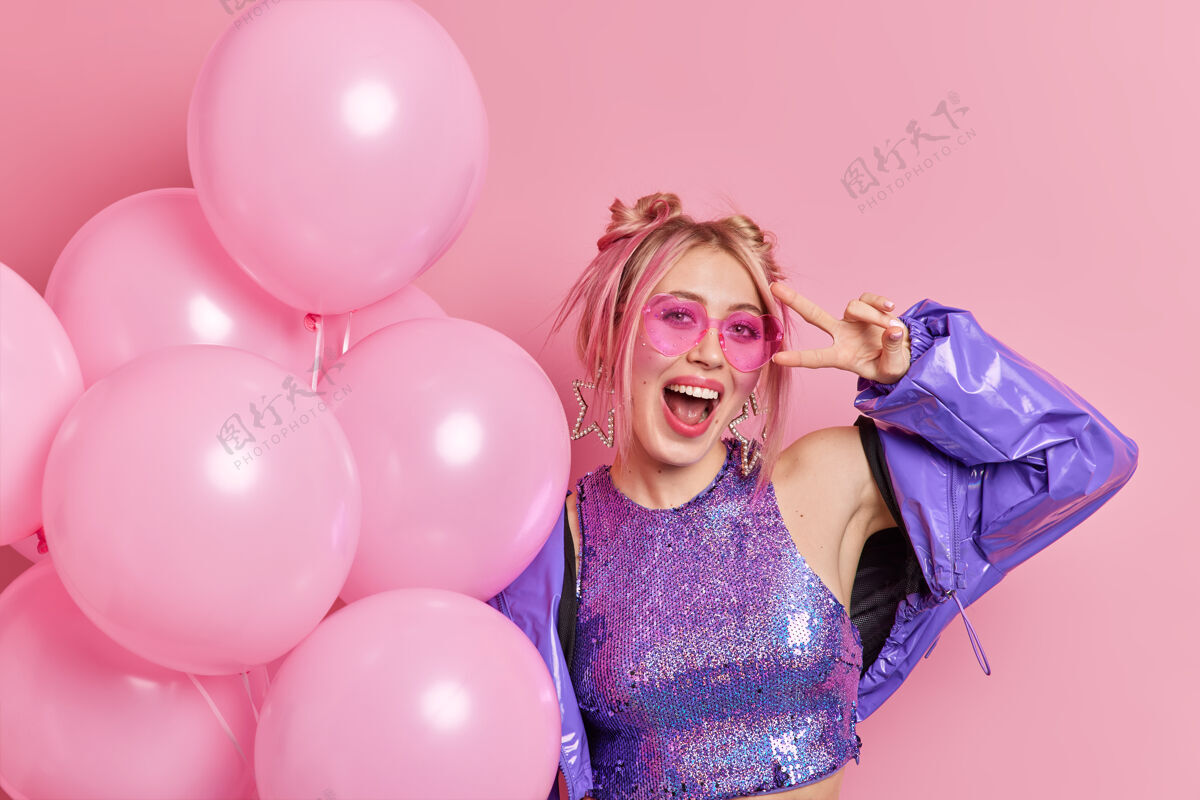 手势无忧无虑的快乐女人的照片生日派对上玩得很开心戴着时髦的太阳镜和紫色夹克欢呼着幸福使和平的姿态拿着一堆充气气球在粉红色的墙上摆姿势和平女性庆典