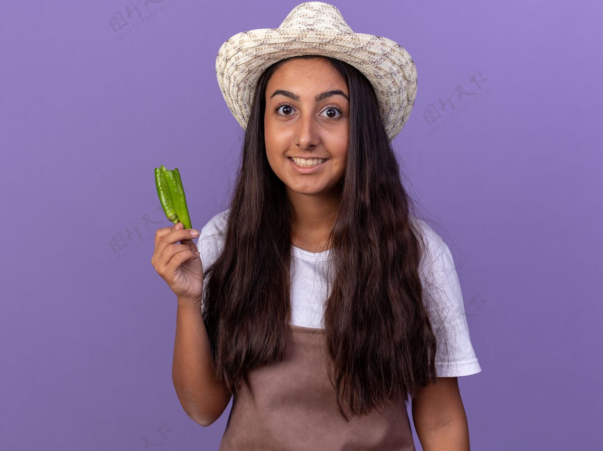 辣椒穿着围裙 戴着夏帽 手里拿着青椒的年轻园丁女孩站在紫色的墙上 又高兴又惊讶年轻帽子围裙