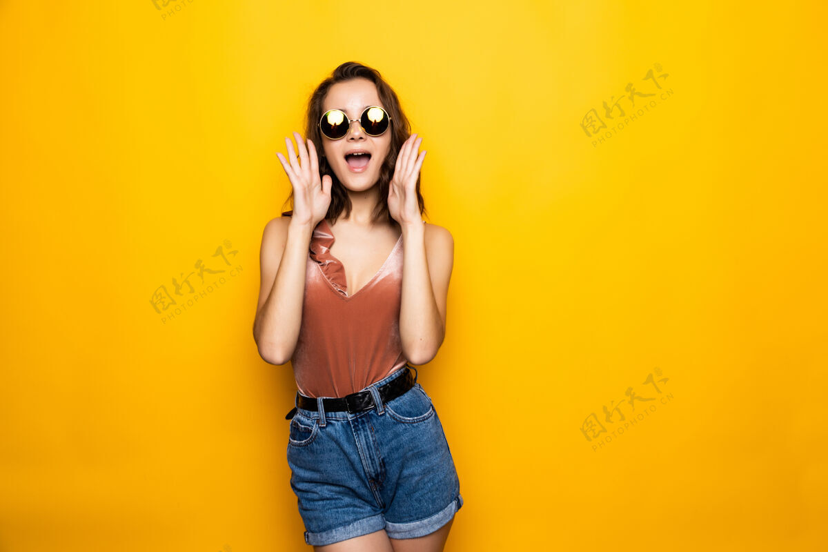 时尚年轻女子墨镜带着惊讶的微笑望向远方 隔离在黄色的墙上惊人奇迹表情
