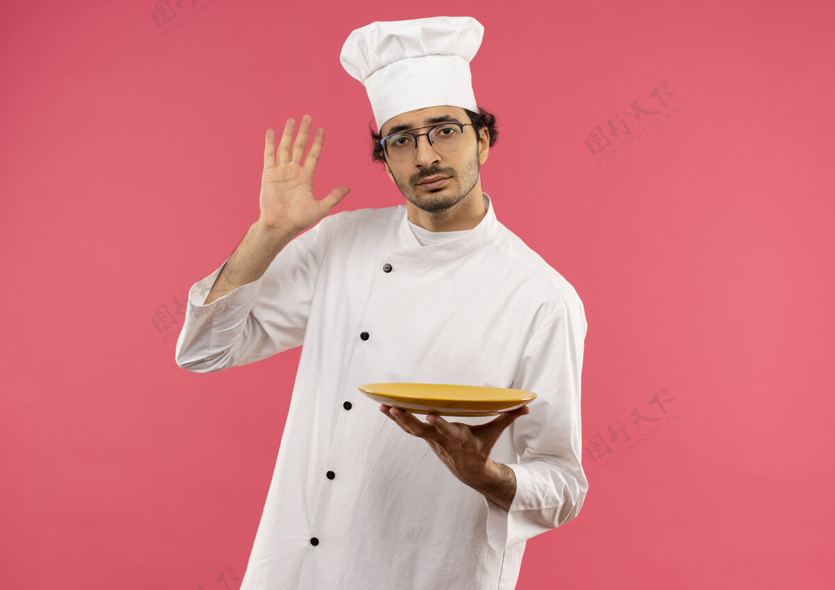 眼镜年轻的男厨师穿着厨师制服 戴着眼镜 拿着盘子举手穿烹饪粉色