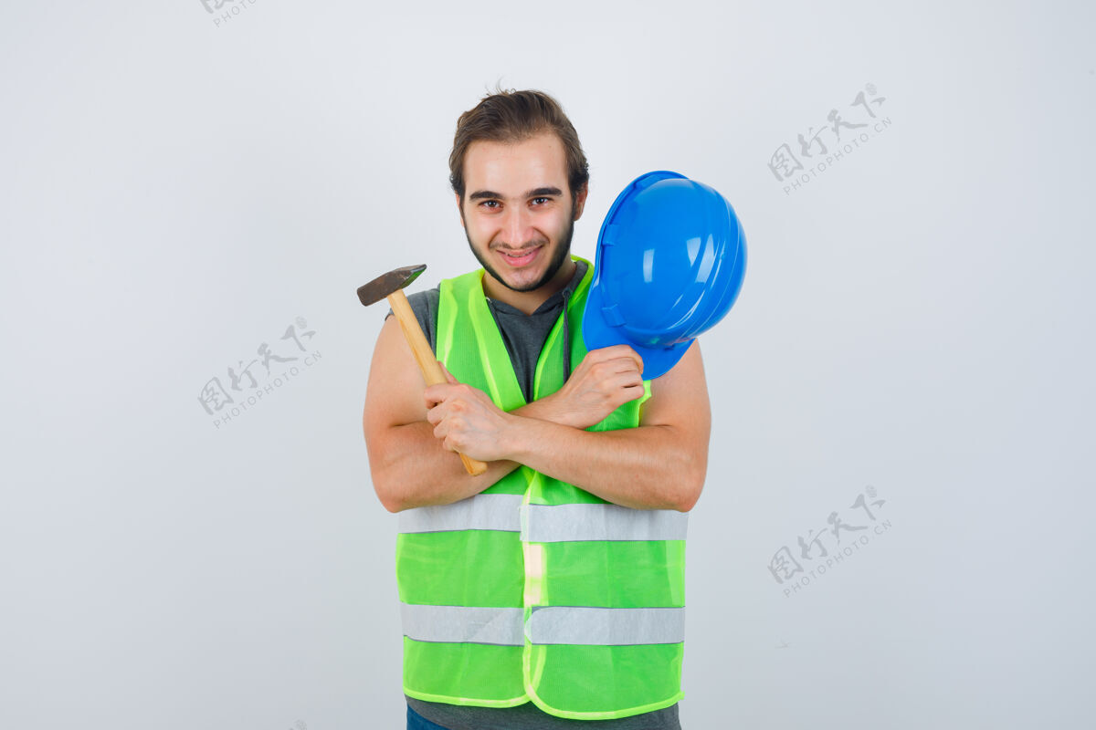 成人年轻的建筑工人手持头盔和锤子 双臂交叉放在胸前 穿着工作服 看上去很高兴正视图男性年轻手臂