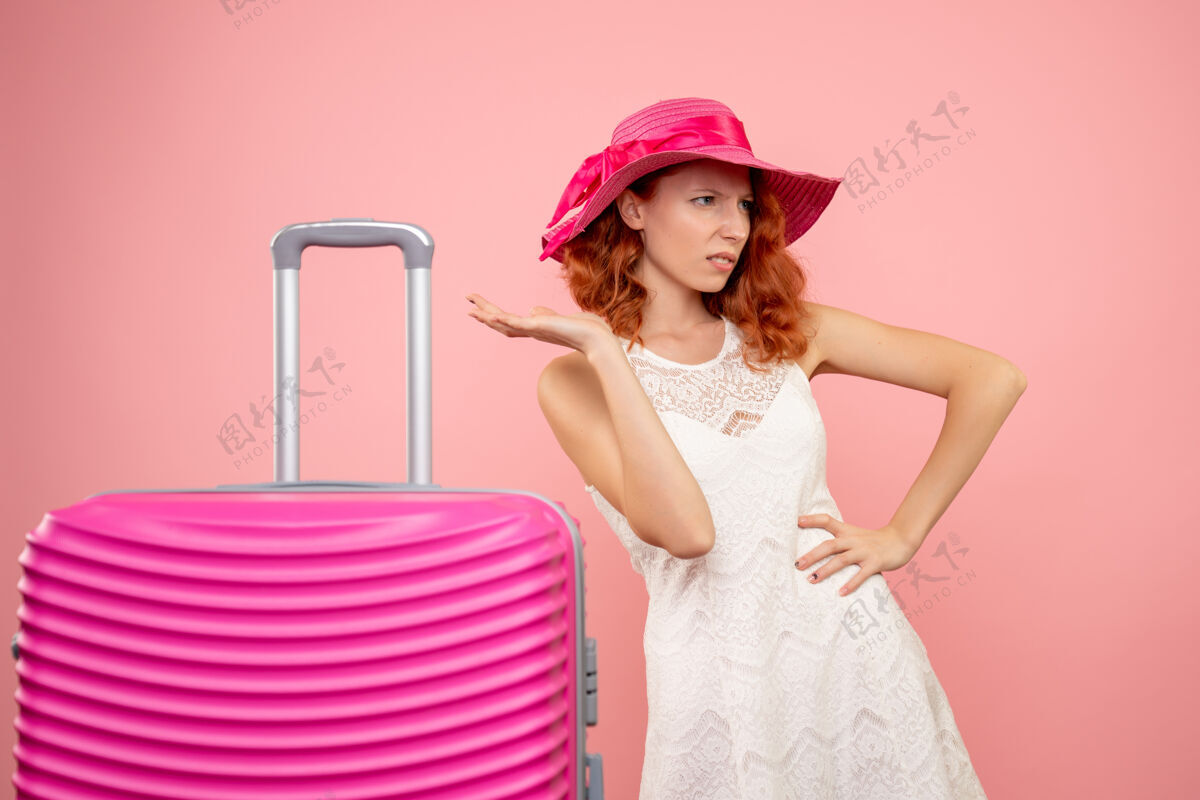 旅行年轻的女性游客与粉红色的帽子和粉红色的墙壁上的包的正面视图肖像快乐假期