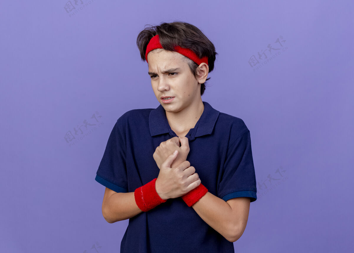 牙套疼痛的年轻英俊的运动男孩戴着头带和戴着牙套的腕带看着一边拿着手腕隔离在紫色墙上的复制空间年轻手腕牙科