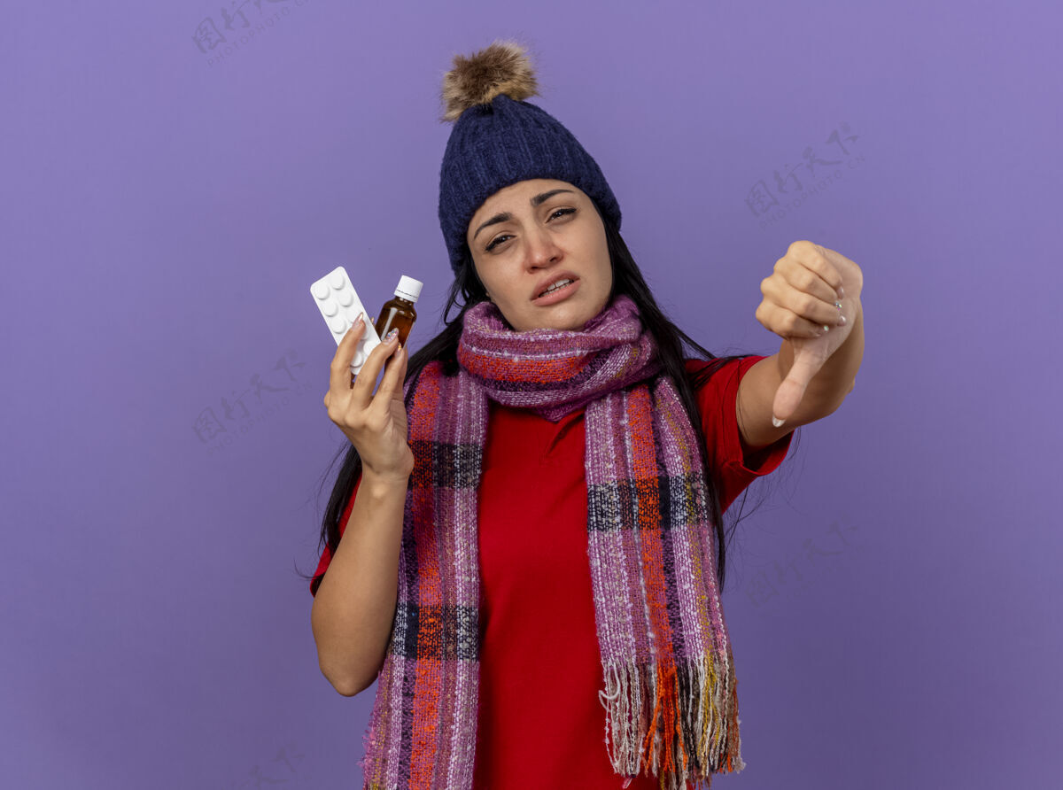 拇指虚弱的年轻白人病女孩戴着冬天的帽子和围巾 手里拿着玻璃药片和一包药片 大拇指朝下 被隔离在紫色的墙上 留着复印空间虚弱显示疾病