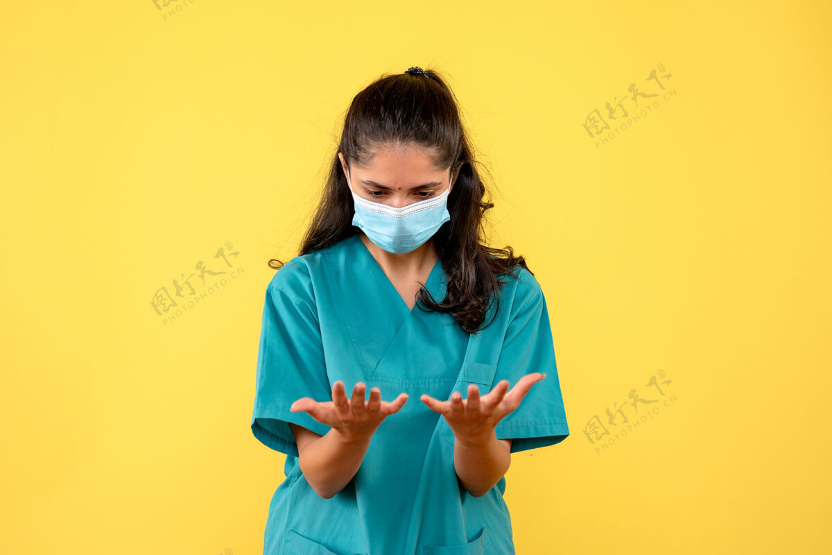 成人戴着医用面罩的漂亮女医生正看着她在黄色墙上的手人手前面