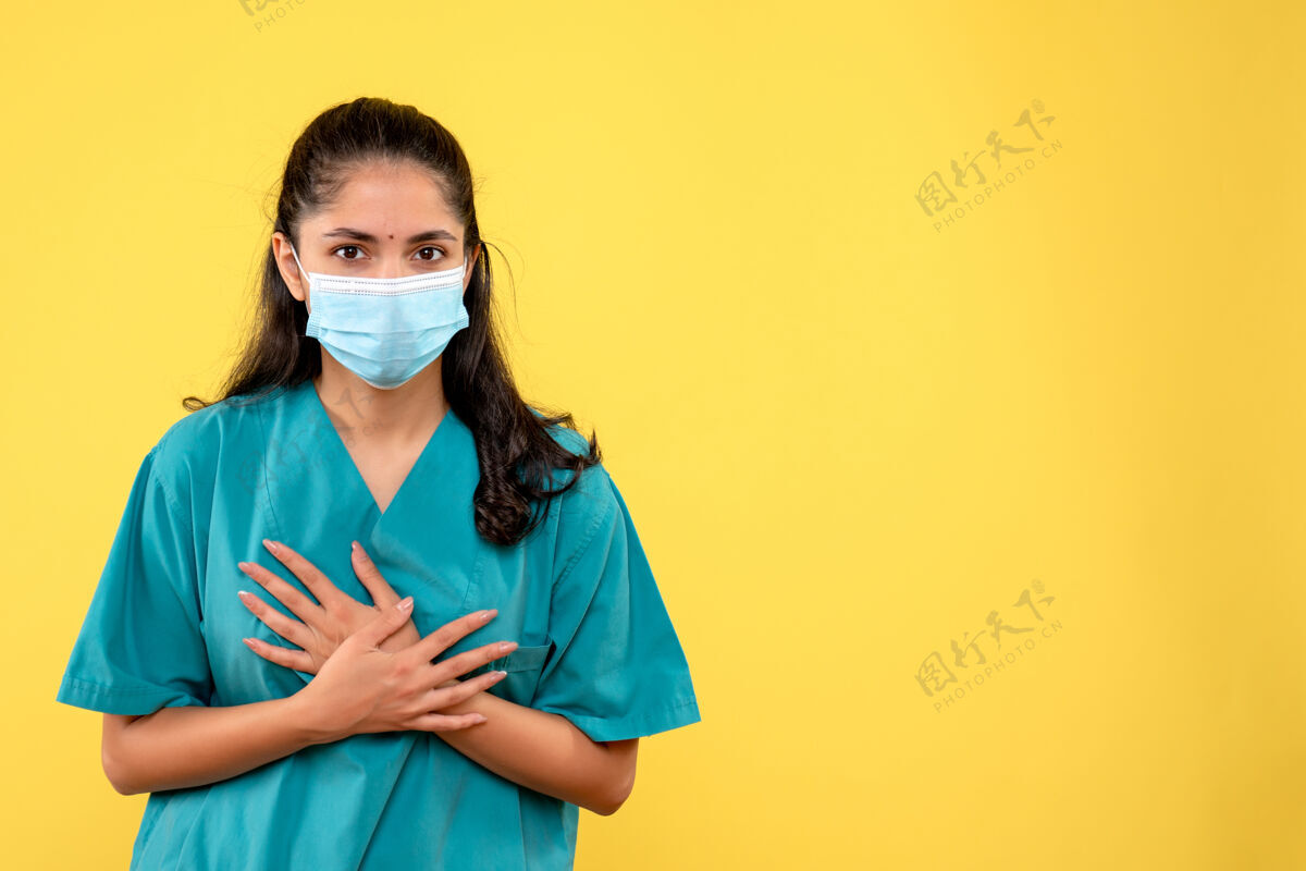 手黄色墙壁上戴着医用口罩的漂亮女医生把手放在胸前的正视图面具医学成人