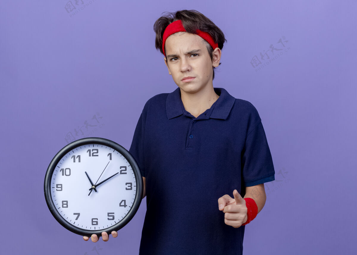 头带皱眉的年轻帅气的运动男孩戴着头带和护腕 戴着牙套 拿着时钟指针 隔离在紫色的墙上 留着复制空间腕带背带穿
