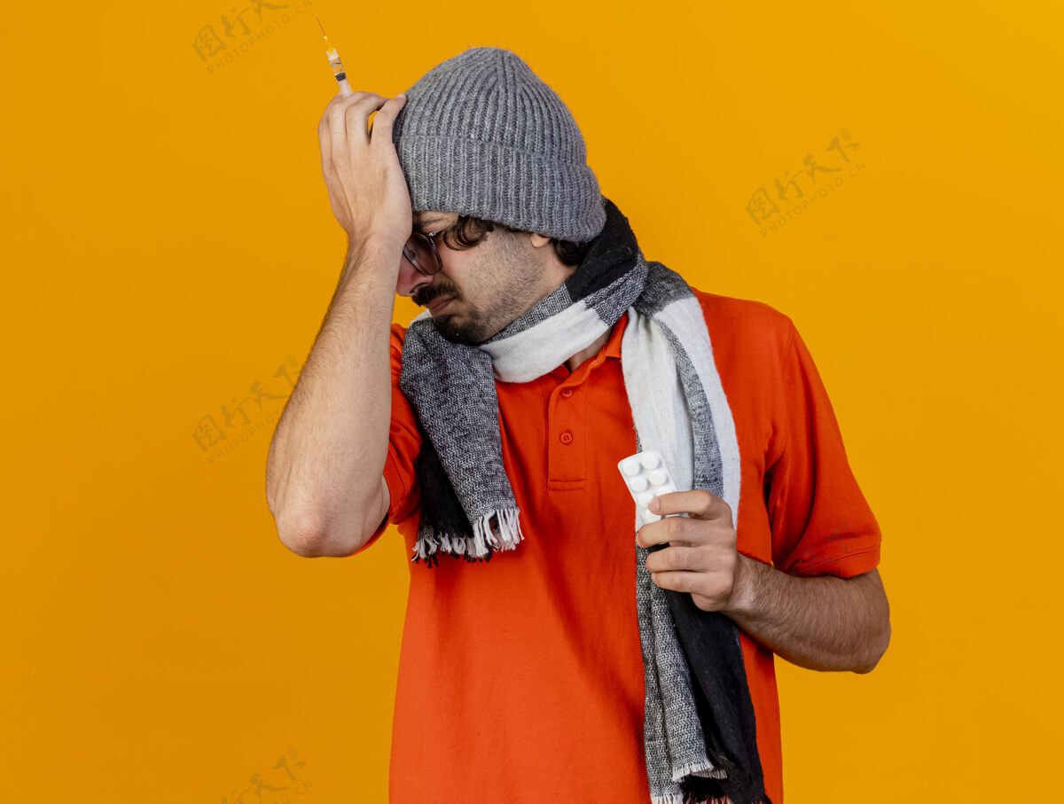 橘子疼痛的年轻白种人 戴着眼镜 戴着冬天的帽子 戴着围巾 手里拿着注射器和一包药片 手放在头上 闭着眼睛 隔离在橙色的墙上 留着复印空间包围巾疼痛