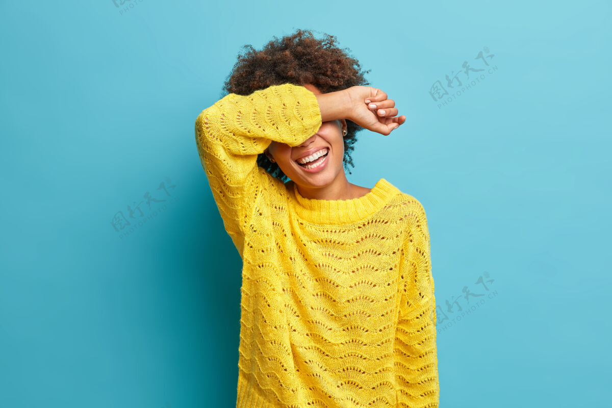 情感一头卷发的正面女人横着镜头真诚地笑着 用胳膊捂着眼睛笑着滑稽的笑话 穿着黄色针织套头衫隔离在蓝色的墙上欢呼隐藏女孩