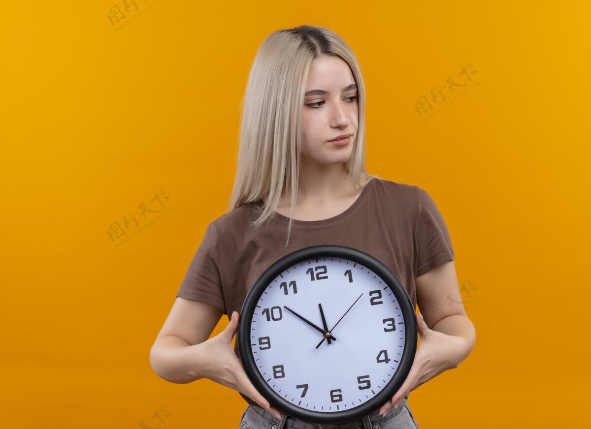 女人年轻的金发女孩拿着时钟看着右边的橙色墙 墙上留着复制空间女孩时钟拿着
