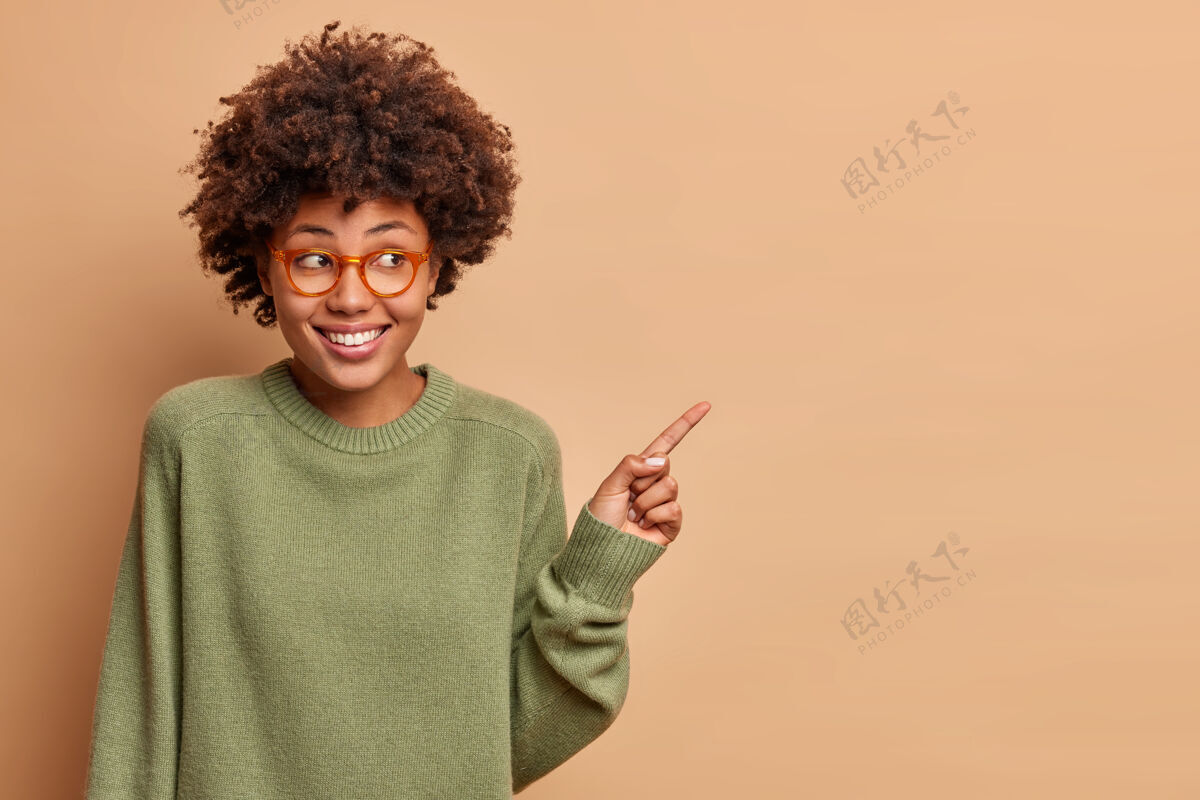 眼镜美丽开朗的女人用食指指着身边有快乐的脸戴眼镜休闲毛衣展示广告内容的复制空间隔离在棕色的墙上正面促销产品