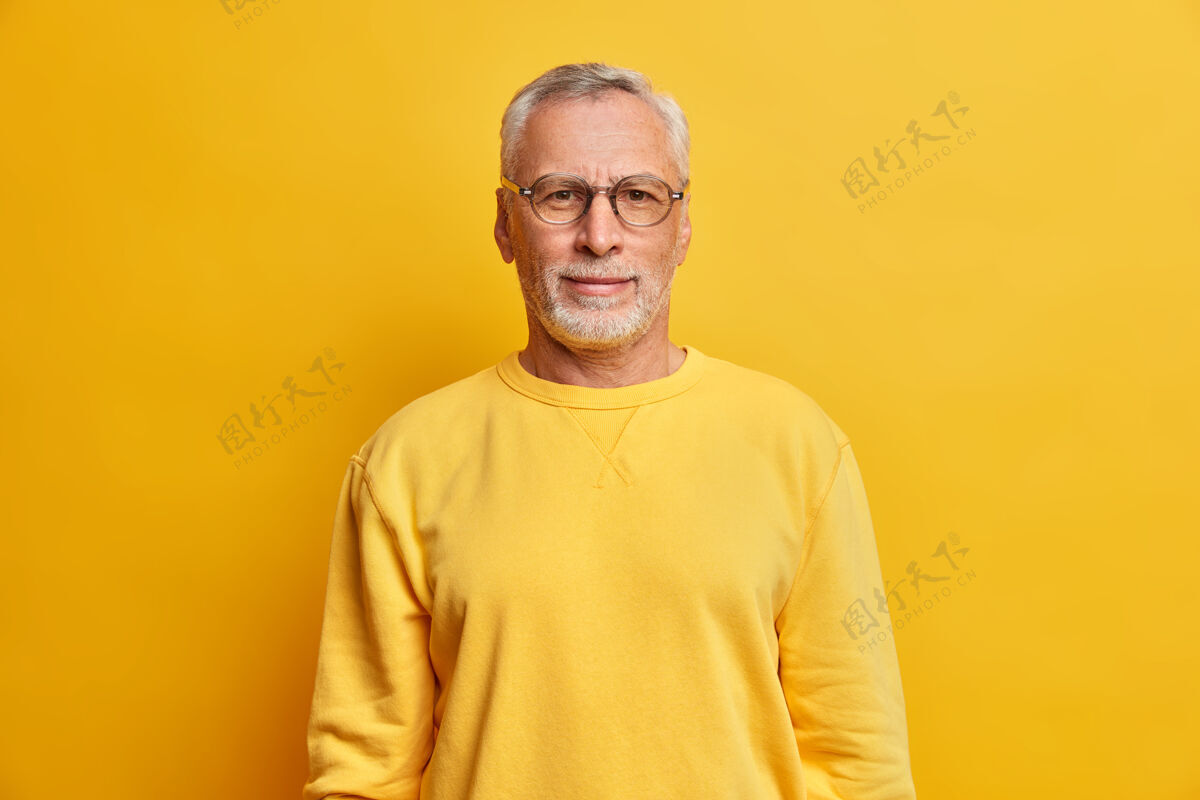 时尚横拍的白发男人带着皱纹戴着眼镜 休闲的黄色套头衫直视前方 在室内摆出满意的表情姿势男性老年人皱眉