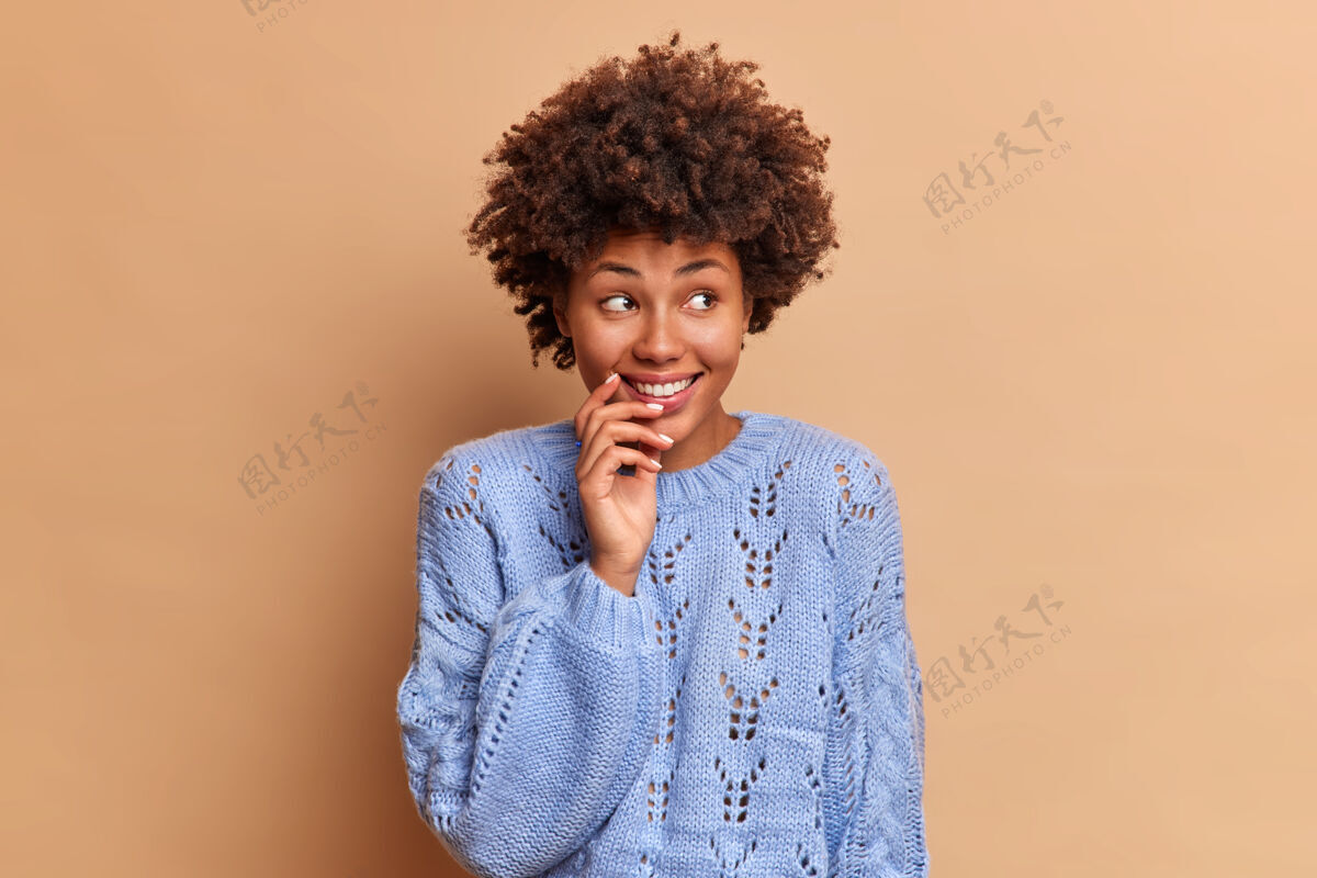 欢乐一个有着非洲式头发 充满梦想 体贴的女人把目光移开 微笑温柔地集中在一边 笑容积极地穿着蓝色套头衫 对着棕色的墙壁摆出姿势有趣沉思正面