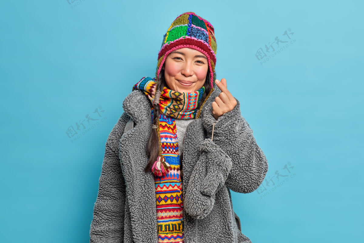 手指可爱的东亚女孩穿着冬装做韩式标志显示迷你们的心表示爱去有散步姿势对蓝色的工作室墙欢呼人围巾