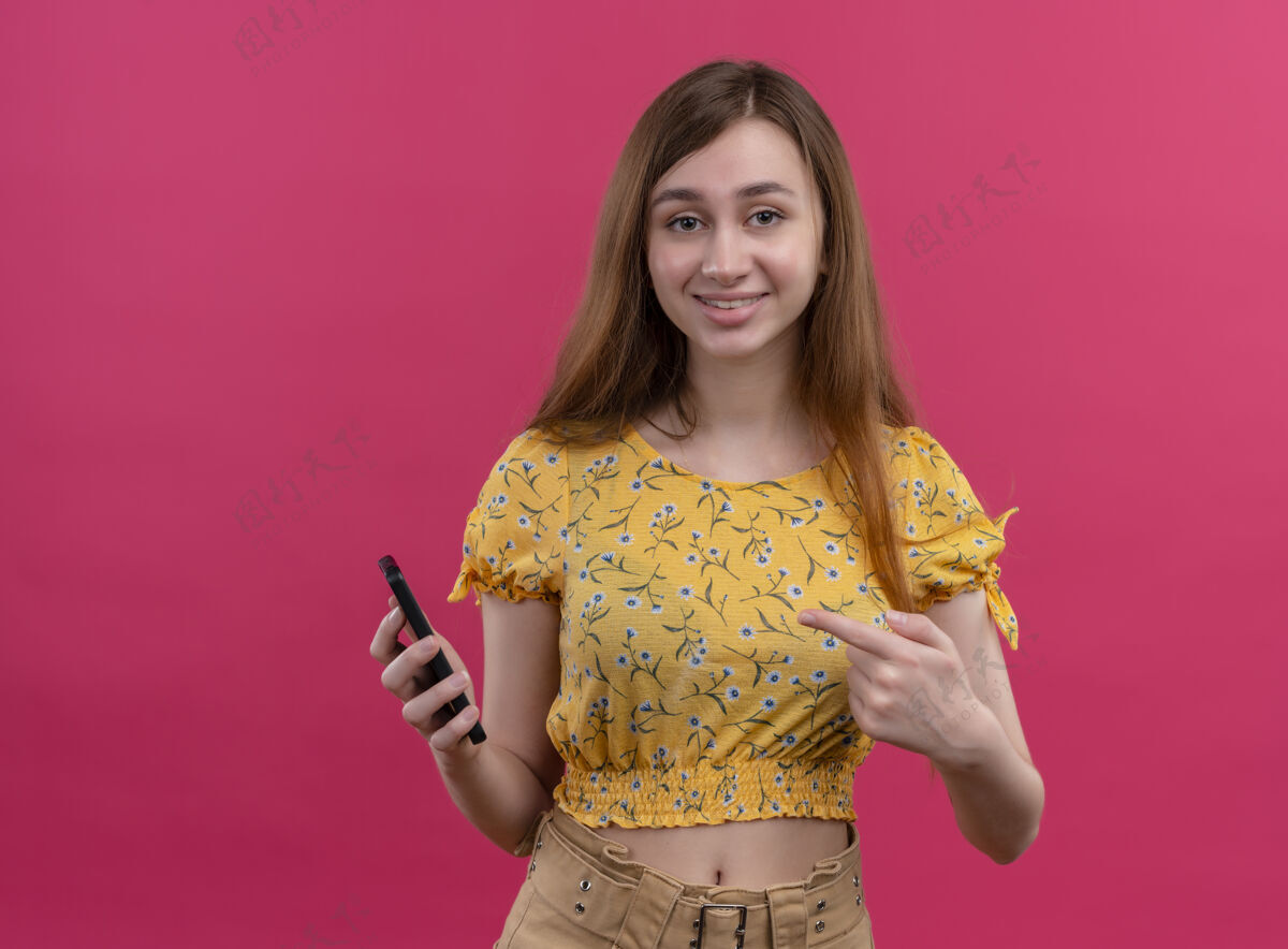 微笑带着微笑的年轻女孩拿着手机 指着隔离的粉色墙壁上有复印空间的手机女孩年轻指着