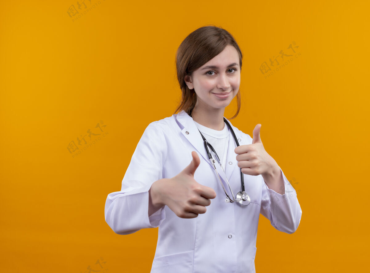 女人微笑的年轻女医生穿着医用长袍和听诊器 在隔离的橙色墙壁上竖起大拇指 留有复印空间听诊器长袍年轻人