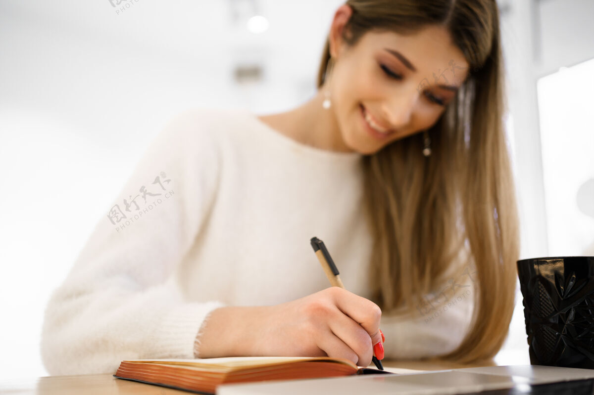 女性微笑美丽的女学生 长发 白色毛衣 在笔记本上写字 同时使用笔记本电脑远程学习远程手机空