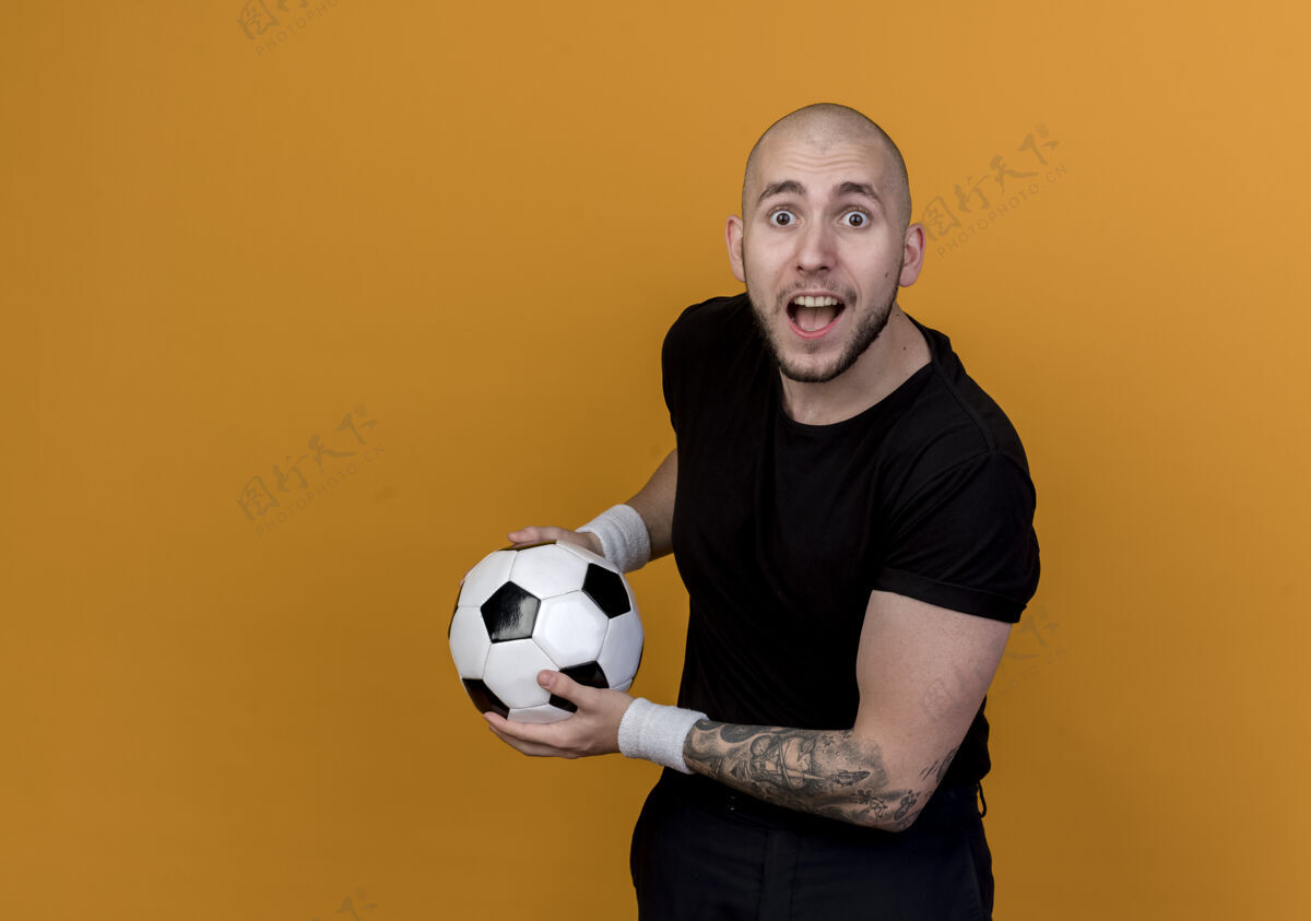 年轻惊讶的年轻人戴着腕带 拿着球 孤立地放在橙色的墙上 留着复制空间男子球惊喜