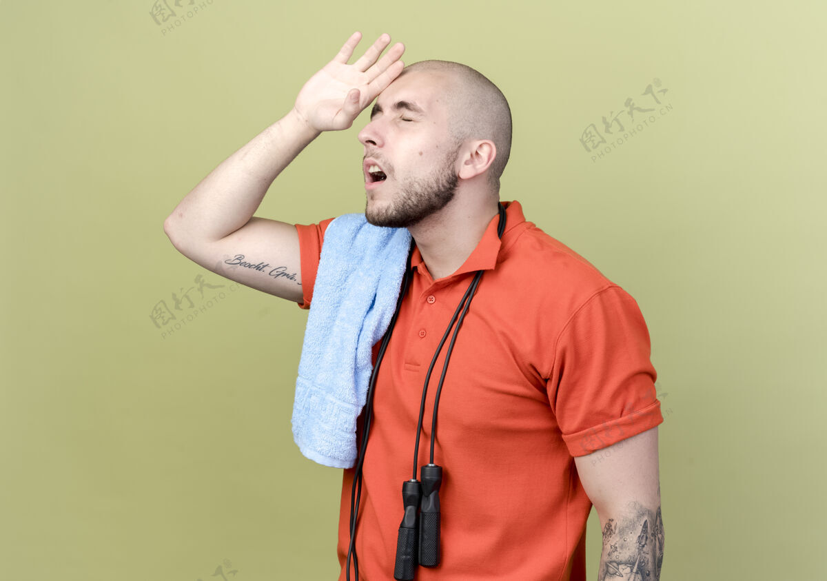 男人闭上眼睛疲惫的年轻运动型男子 肩上拿着毛巾和跳绳 手放在额头上 隔离在橄榄绿的墙上手疲倦前额