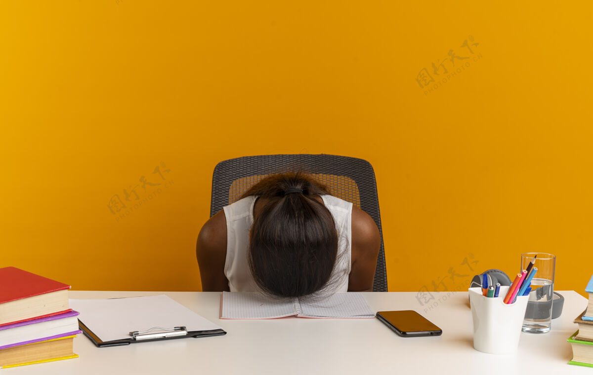 书桌低着头的年轻女学生坐在书桌旁 把学习工具孤立在橙色的墙上头年轻低一点