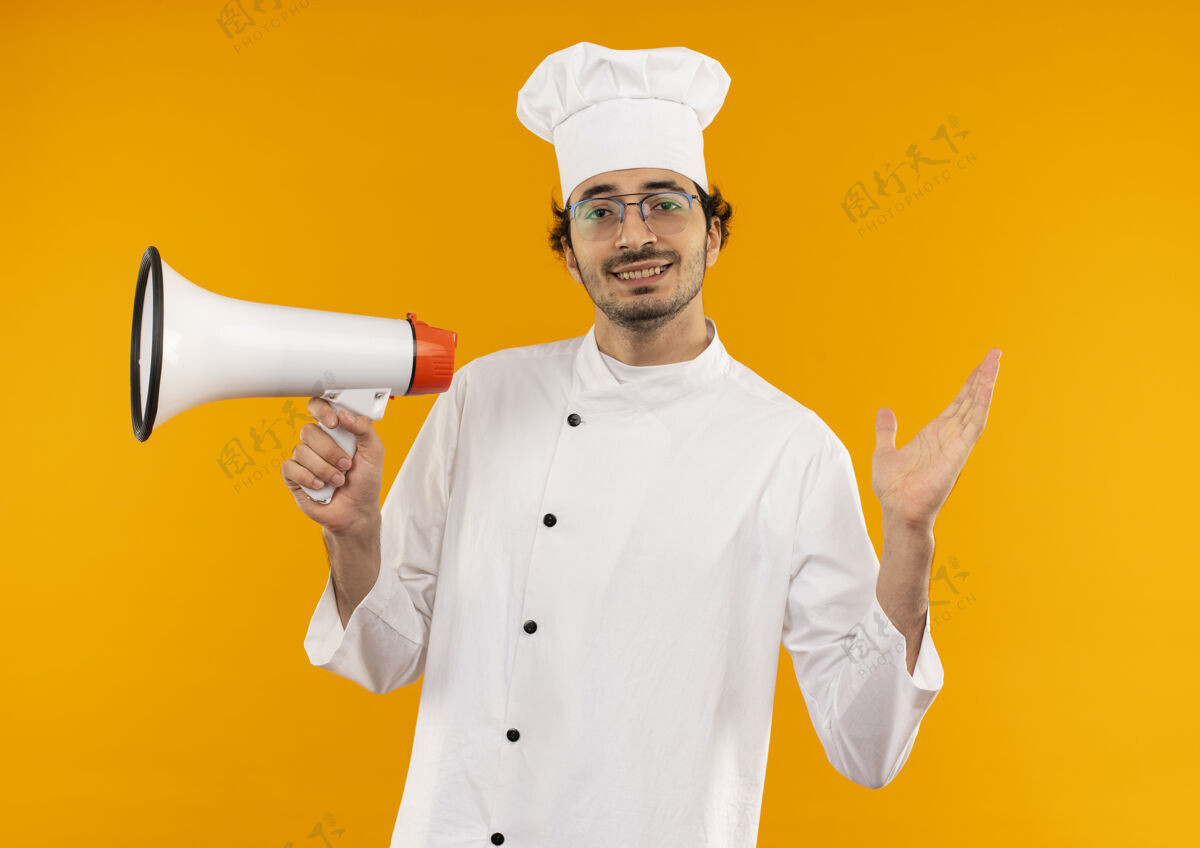 厨师面带微笑的年轻男厨师身穿厨师制服 戴着眼镜 手持扩音器 将手孤立地摊开在黄色的墙上传播男性手