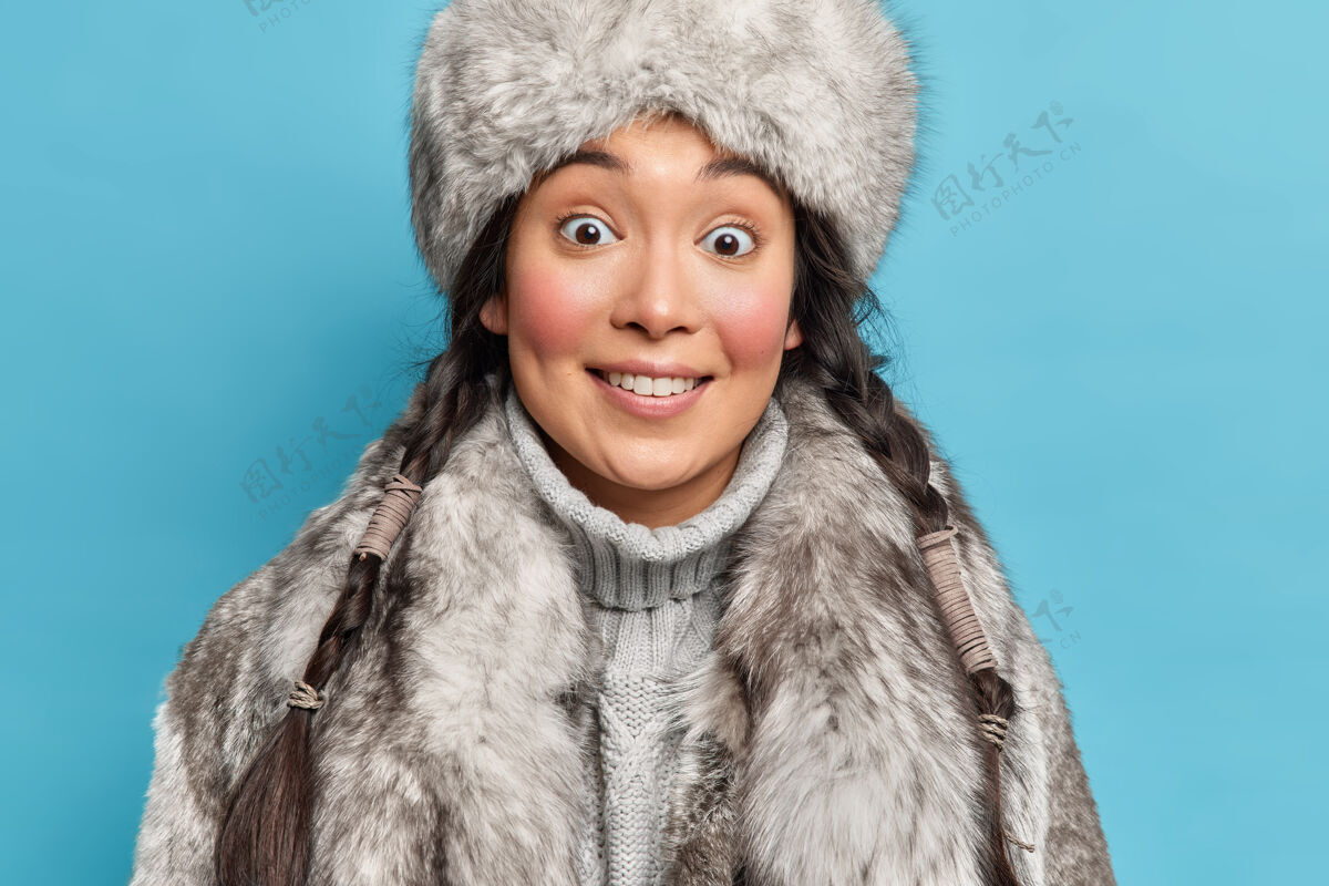 霜冻一位留着两条辫子 身穿毛灰色外套的亚洲妇女的特写镜头 微笑着愉快地生活在隔离在蓝色墙壁上的北极地区民族手套头饰