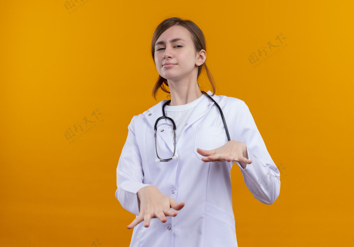 伸展穿着医用长袍和听诊器的年轻女医生在隔离的橙色墙壁上伸展双手 并留有复印空间穿女人医学