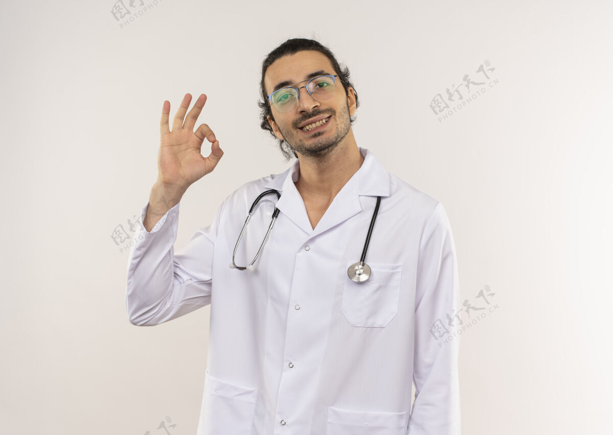 微笑微笑的年轻男性医生 戴着眼镜 穿着白色长袍 听诊器 在隔离的白色墙壁上显示出良好的姿势 并留有复印空间听诊器年轻展示