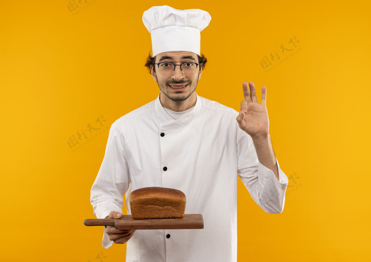好的面带微笑的年轻男厨师 身穿厨师制服 戴着眼镜 面包放在砧板上 黄色的墙上显示奥基被隔离了厨师男性年轻