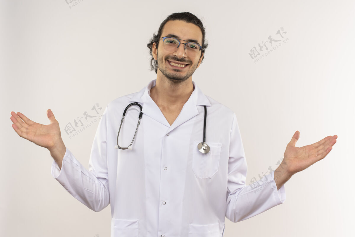 男性微笑着的年轻男医生戴着眼镜 穿着白色长袍 带着听诊器 在隔离的白色墙壁上摊开双手 留着复印空间年轻传播手