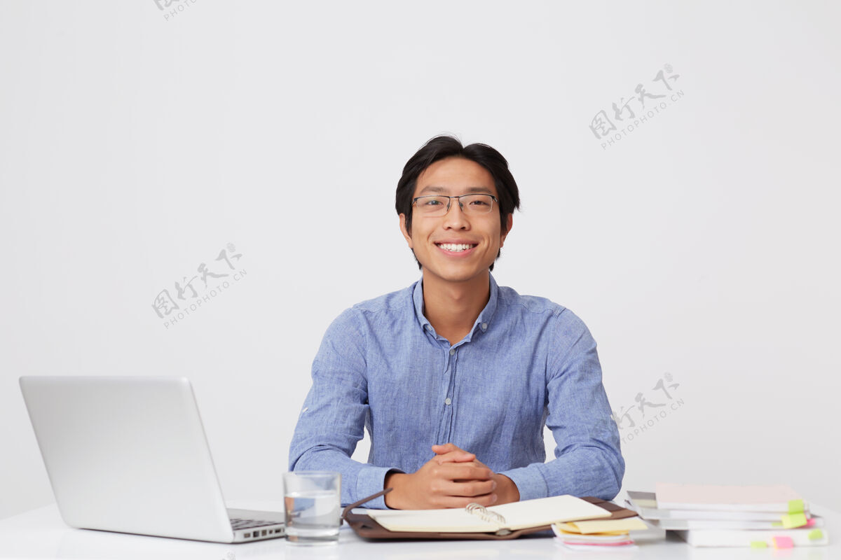 坐着的人快乐英俊的亚洲年轻商人戴着眼镜 穿着蓝色衬衫 坐在桌边 笔记本电脑盖在白墙上高兴学生眼镜