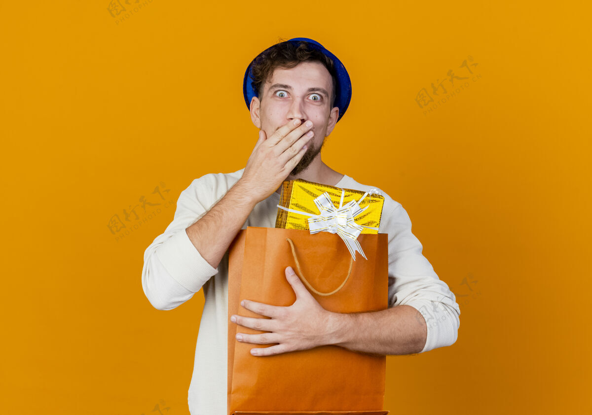 手惊讶年轻英俊的斯拉夫党人戴着党的帽子拿着礼品盒在纸袋里看着相机保持手放在嘴上橙色的背景与复制空间隔离帅哥盒子拿着