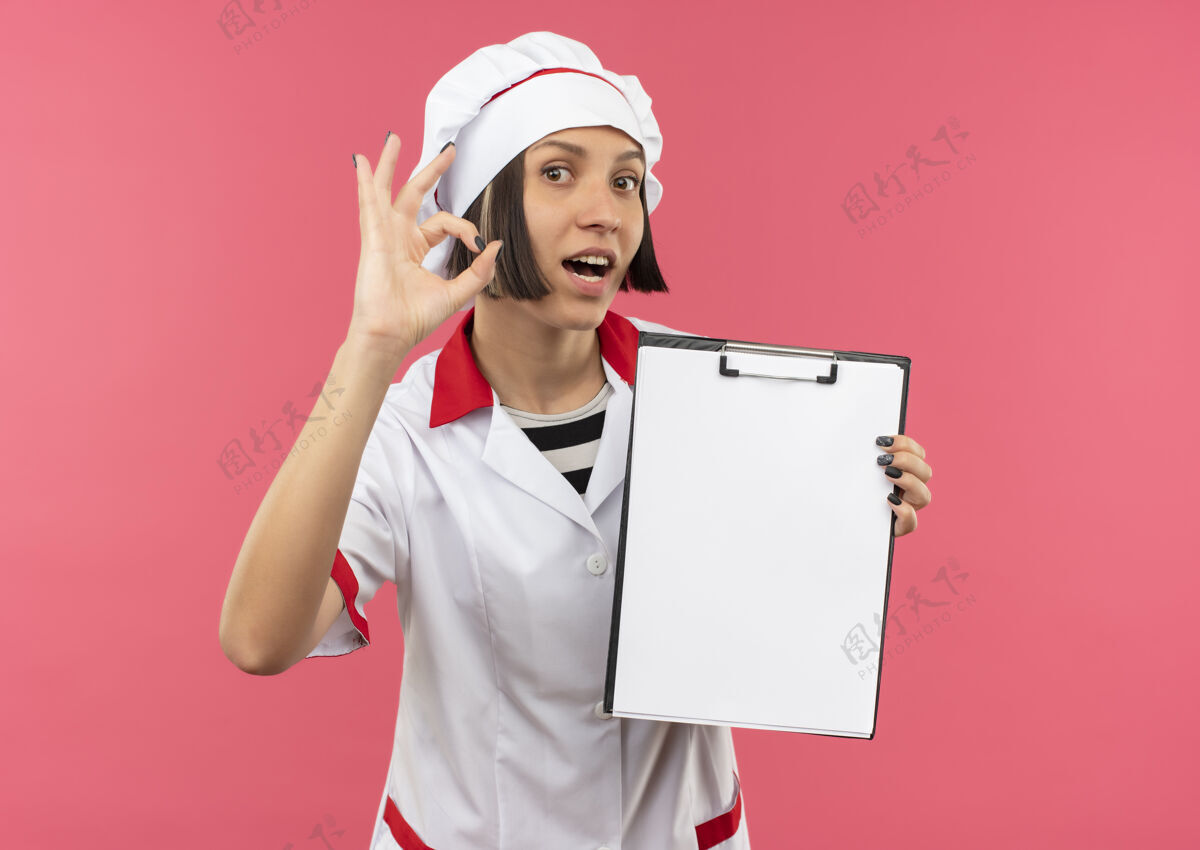 烹饪令人印象深刻的年轻女厨师在厨师制服举行剪贴板和做ok标志孤立的粉红色剪贴板好印象
