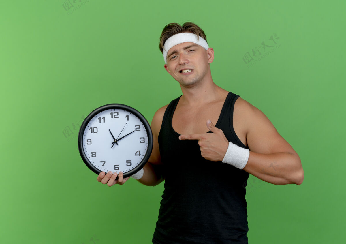 自信自信的年轻帅气的运动型男人戴着头带和腕带拿着并指着绿色的时钟腕带绿色头带