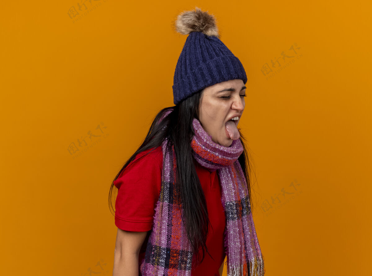 年轻愤怒的年轻白种人生病的女孩戴着冬天的帽子和围巾站在侧视图显示舌头闭着眼睛孤立在橙色背景与复制空间站着帽子舌头