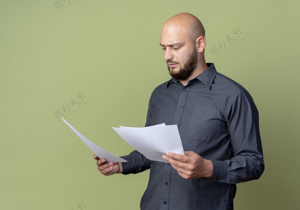 年轻专注的年轻秃头呼叫中心男子拿着和橄榄绿孤立的文件看文件橄榄呼叫
