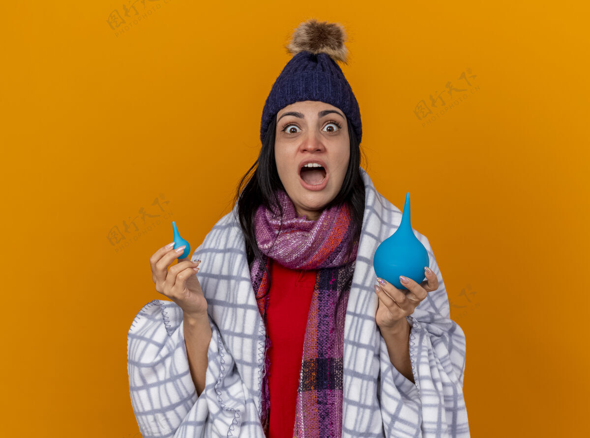 拿着印象深刻的年轻白种人生病的女孩戴着冬天的帽子和围巾包裹在格子布拿着灌肠看着相机孤立的橙色背景与复制空间年轻围巾疾病