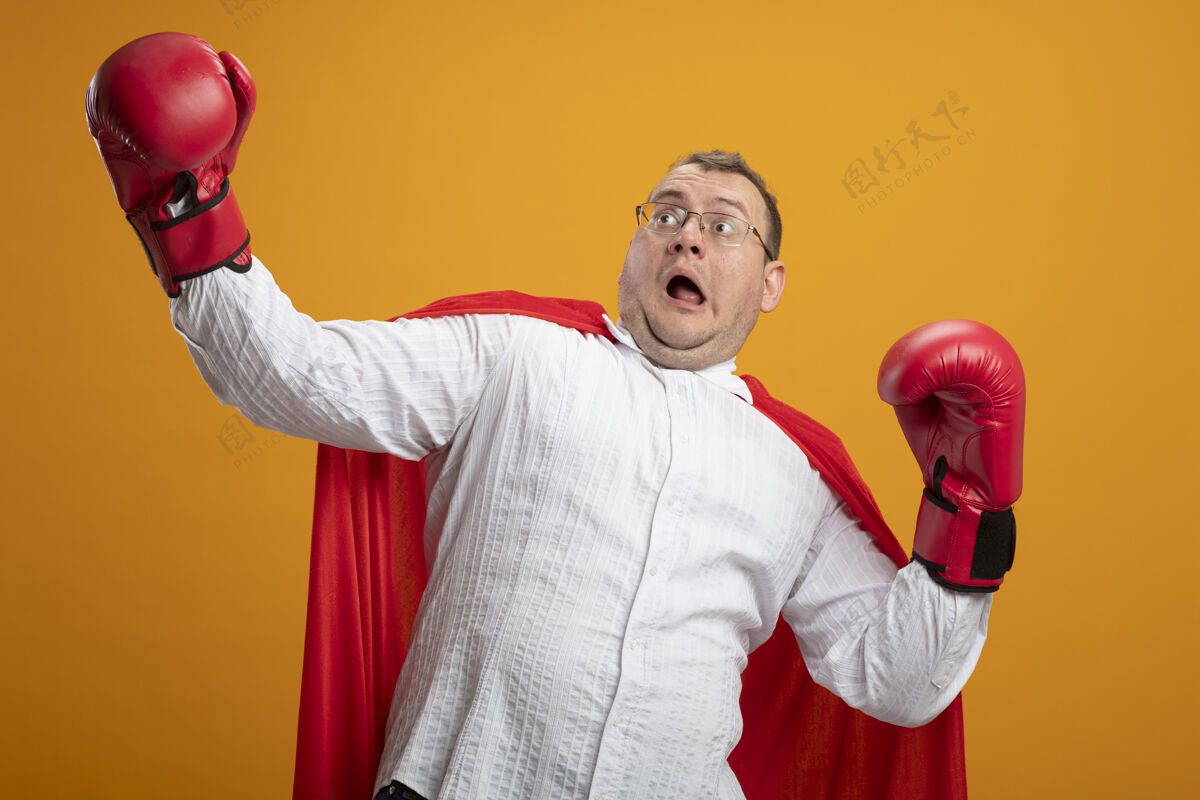 超级英雄被吓坏了的成年斯拉夫超级英雄 穿着红色斗篷 戴着眼镜和箱手套 看着一边 手在空气中被隔离在橙色背景上保持眼镜斗篷